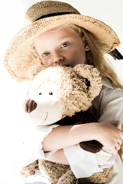 beautiful child in straw hat hugging teddy bear and looking at camera on white  - Photo, Image