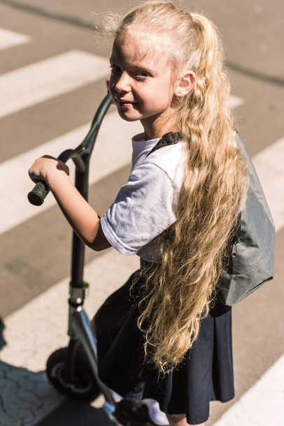 high angle view of beautiful little schoolgirl with long curly hair riding scooter and looking at camera - Foto, imagen