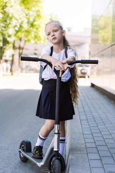 cute pensive schoolchild with backpack riding scooter and looking away on street - Foto, Bild