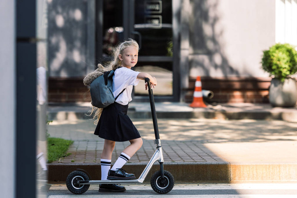 adorable schoolgirl with backpack and teddy bear riding scooter and looking at camera on street - Zdjęcie, obraz