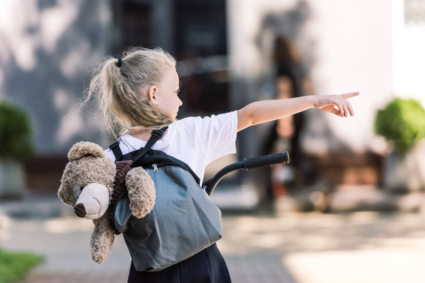 back view of adorable little schoolgirl with backpack and teddy bear riding scooter and pointing away with finger on street - Photo, Image