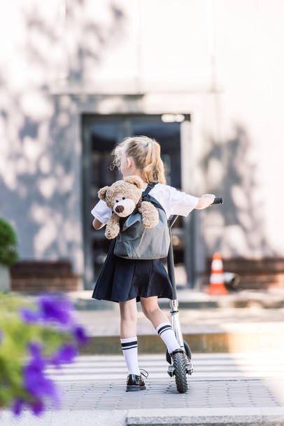 back view of adorable little schoolgirl with backpack and teddy bear riding scooter on street   - Photo, Image