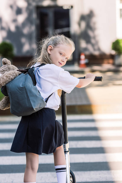 back view of adorable little schoolgirl with backpack and teddy bear riding scooter and looking at camera - 写真・画像