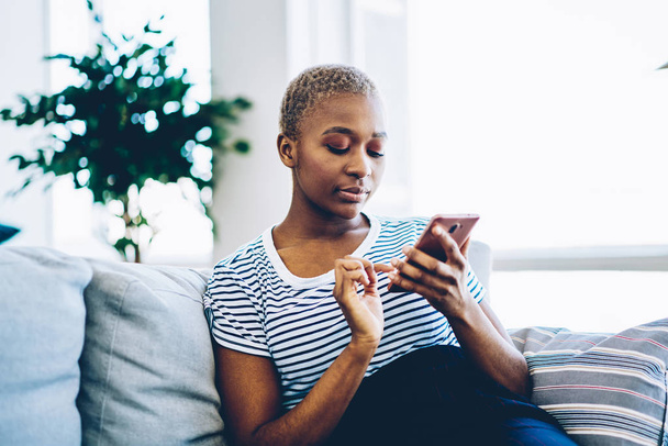 Pensive african american hipster girl making banking via application on smartphone while sitting on sofa at home, serious dark skinned woman checking new feed on social networks using mobile phon - Fotografie, Obrázek