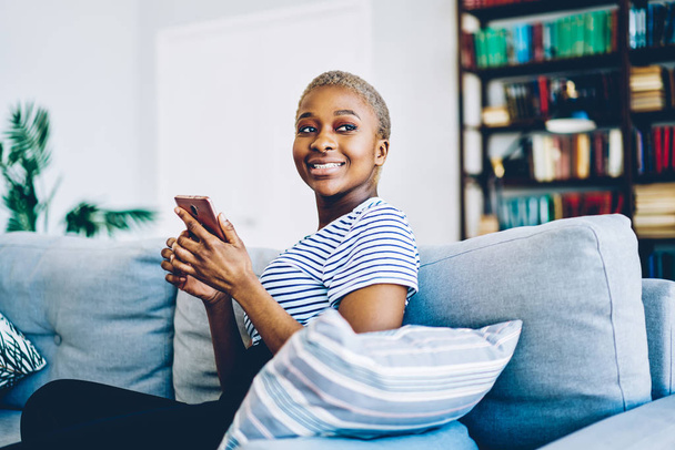 Cheerful african american woman satisfied with wireless internet at home connecting smartphone for chatting and blogging,happy dark skinned hipster girl using mobile phone for sending messag - Fotografie, Obrázek