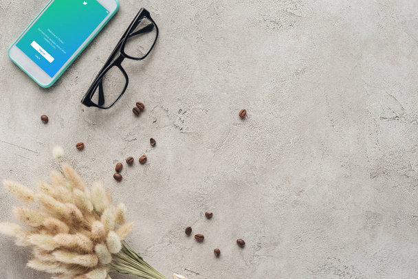 top view of smartphone with twitter app on screen with eyeglasses, spilled coffee beans and lagurus ovatus bouquet on concrete surface - Photo, image