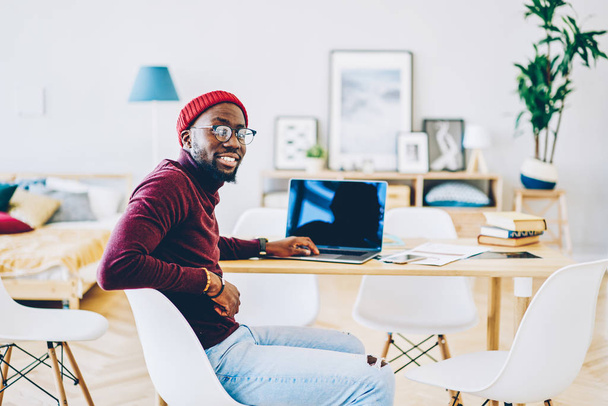 Portrait of cheerful african american male freelancer sitting at desktop with laptop computer satisfied with successful online startup, smiling dark skinned hipster guy looking at camera update ap - Foto, Imagem