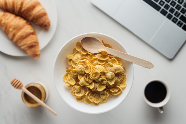 Blick von oben auf Schüssel mit Cornflakes mit leckerem Kaffee, Croissants und Laptop auf weiß - Foto, Bild