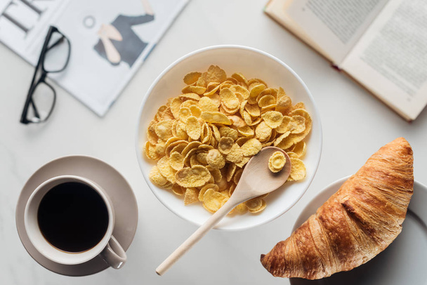 vista superior del tazón de desayuno de cereales secos con taza de café y croissant en la superficie blanca con periódico y libro
 - Foto, imagen