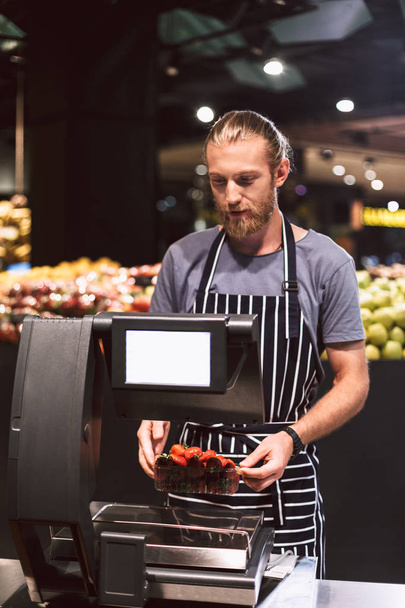 Jonge verkoper in schort met een gewicht van aardbeien op schaal in moderne supermarkt - Foto, afbeelding