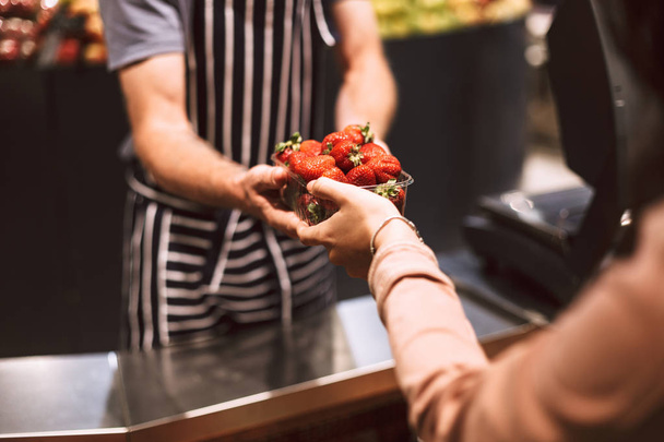 Εσωτερικη πωλητής στο ριγέ ποδιά πίσω από το αντίθετο που δίνει τις φράουλες σε πελάτη στην σύγχρονη υπεραγορά  - Φωτογραφία, εικόνα