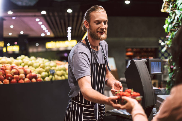 Smiling salesman in striped apron behind counter dreamily giving strawberries to customer in modern supermarket - Photo, Image