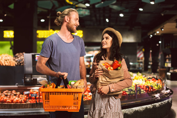 junger lächelnder Kerl und hübsches Mädchen, die sich glücklich anschauen, mit Korb und Papiertüte voller Produkte im modernen Supermarkt - Foto, Bild