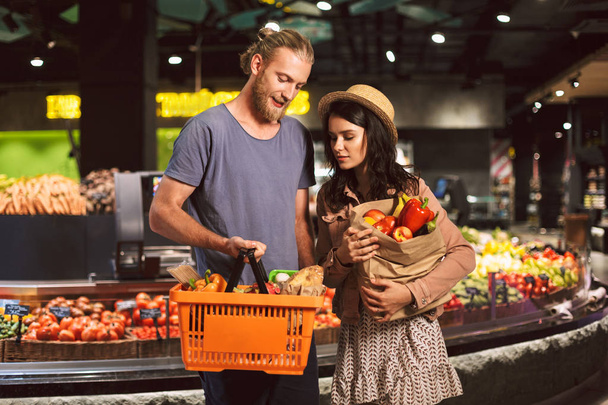 Junge Kerle und Mädchen schauen verträumt auf Korb und Papiertüte voller Produkte im modernen Supermarkt - Foto, Bild