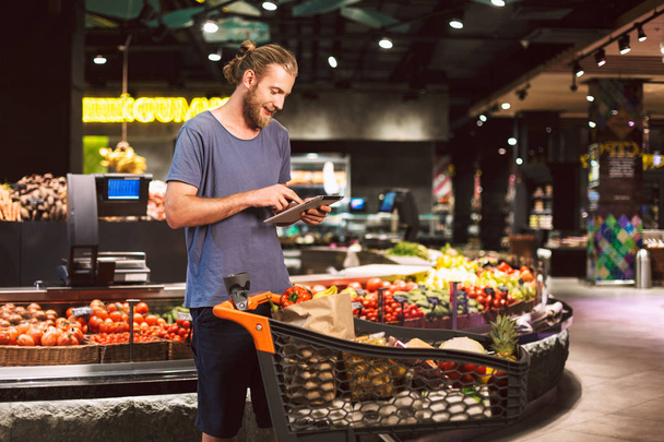 Joven hombre sonriente con carro lleno de productos felizmente usando la tableta para leer la lista de compras en el supermercado moderno
 - Foto, imagen