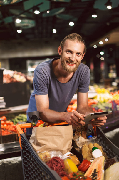 Молодий усміхнений хлопець, спираючись на візок, сповнений продуктів радісно дивиться в камеру, тримаючи планшет в руці в сучасному супермаркеті
 - Фото, зображення