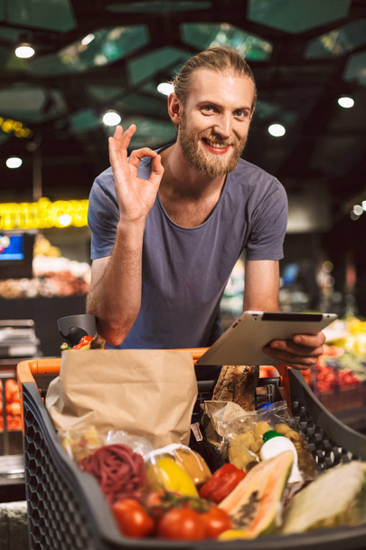 Mladý, usměvavý muž s vozíkem plné potravy šťastně dívá za zavřenými zobrazeno pořádku gesto s tablety v ruce v moderním supermarketu - Fotografie, Obrázek