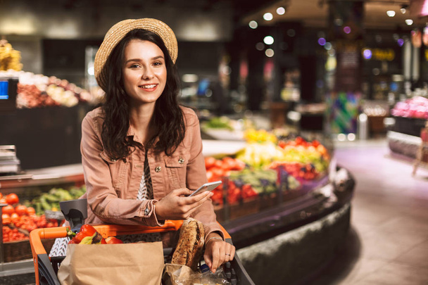 Mooi lachende meisje in hoed leunend op trolley koffer vol producten gelukkig kijken in de camera met mobiele telefoon in de hand in moderne supermarkt - Foto, afbeelding