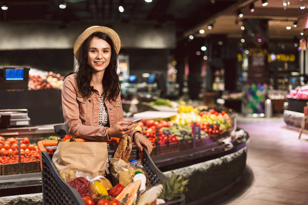 Menina muito sorridente em chapéu com carrinho cheio de produtos alegremente olhando na câmera com celular na mão no supermercado moderno
 - Foto, Imagem