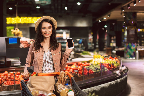 hübsches lächelndes Mädchen mit Hut und Einkaufswagen voller Produkte, das freudig in die Kamera schaut und das neue Handy in einem modernen Supermarkt zeigt - Foto, Bild