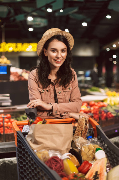 Kaunis hymyilevä tyttö hattu nojaten vaunun täynnä tuotteita iloisesti etsivät kameran viettää aikaa moderni supermarket
 - Valokuva, kuva