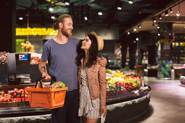 Nuori kaunis pari onnellisesti katsomalla toisiaan kori täynnä tuotteita viettää aikaa yhdessä moderni supermarket
 - Valokuva, kuva