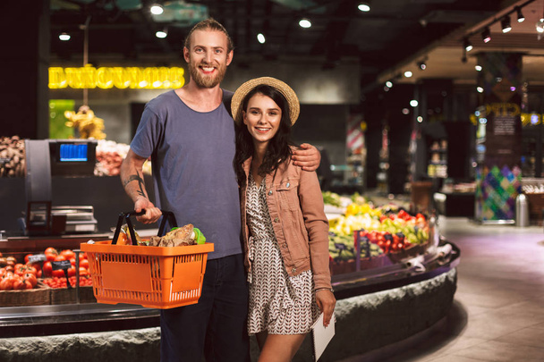 Nuori kaunis pari onnellisena kameran edessä kori täynnä tuotteita viettää aikaa yhdessä moderni supermarket
 - Valokuva, kuva