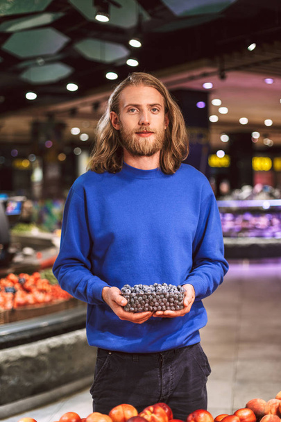 Chico joven en suéter azul oscuro mirando soñadoramente en la cámara con arándanos en la mano en el supermercado moderno
 - Foto, imagen