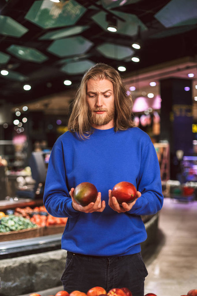 Jeune homme en pull bleu foncé regardant attentivement les mangues en main dans un supermarché moderne
 - Photo, image
