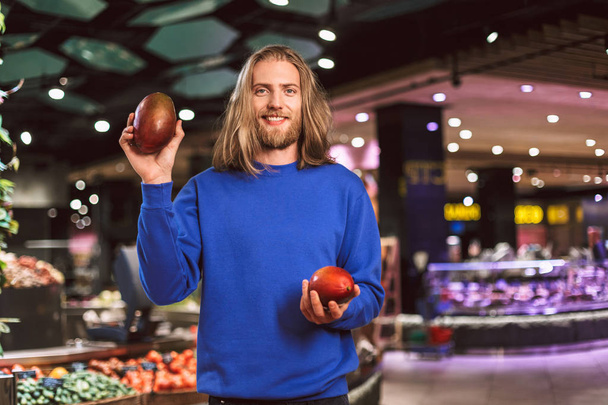 Junger lächelnder Kerl in dunkelblauem Pullover, der freudig in die Kamera blickt und im modernen Supermarkt Mango in den Händen hält - Foto, Bild