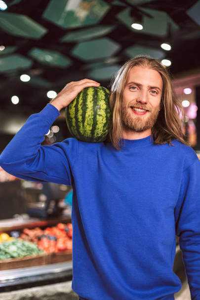 Portrait of joyful guy in dark blue sweater happily looking in camera holding watermelon on shoulder in modern supermarket - Foto, immagini