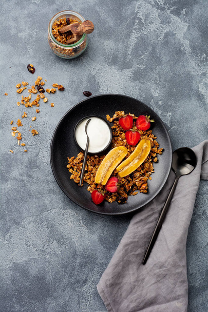 Oatmeal granola with yoghurt, fresh strawberries and banana, chia seeds, sunflower and honey in black ceramic plate on dark background. Top view. - Photo, image