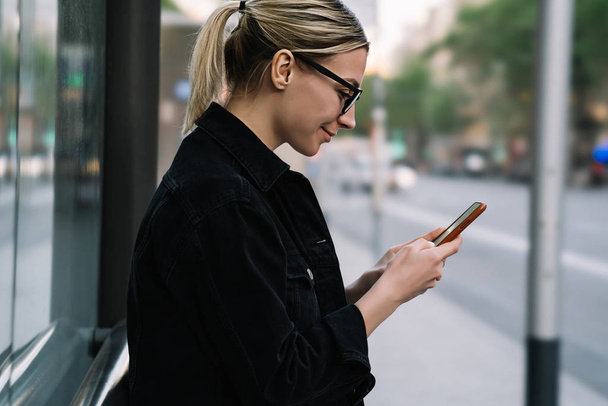 Side view of cheerful young woman blogger laughing while chatting online in social networks on modern smartphone using 4G internet.Positive hipster girl holding cellular and reading news on website - Photo, Image
