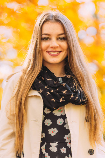 Beautiful blonde young woman in nice white coat and scarf. Posing on golden autumn background. Autumn woman having fun at the park and smiling. Young woman portrait in autumn color. - Photo, image