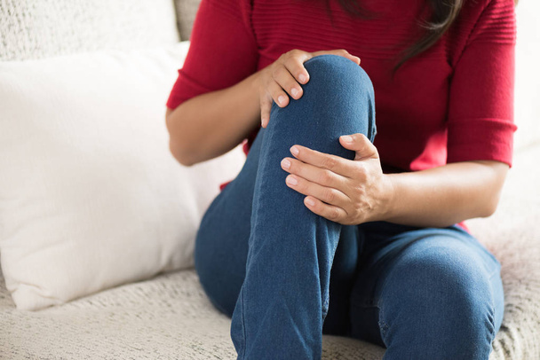 Mujer joven de primer plano sentada en el sofá y sintiendo dolor de rodilla y se masajea la rodilla en casa. Salud y concepto médico
. - Foto, imagen