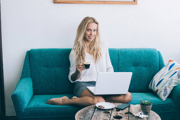 Successful hipster girl sitting on couch in living room using laptop device and wireless connection while looking at camera, portrait of positive cheerful woman holding cup of hot coffee in home - Fotografie, Obrázek
