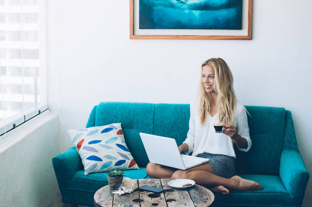 Cheerful hipster girl sitting on cosy couch with laptop computer in home interior working on freelance successfully, positive woman enjoying coffee time while using wireless connection indoors - Photo, image