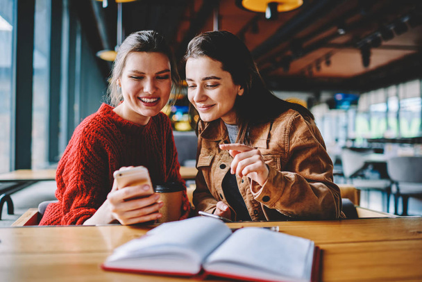 Happy caucasian hipster girls reading text message from friend using telephone device for communication, positive women spending free time with technology at college campus in break of studying - Фото, изображение