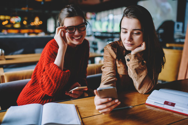 Attractive caucasian hipster girl open bluetooth instruction on mobile phone while happy female friend in spectacles sitting near looking at her telephone and waiting for sharing information - 写真・画像