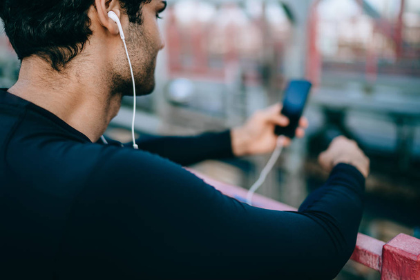Back view of young sportsman making little break in training outdoors and searching new songs for motivation, active male athlete listening music via electronic headphones and mobile application - Foto, immagini