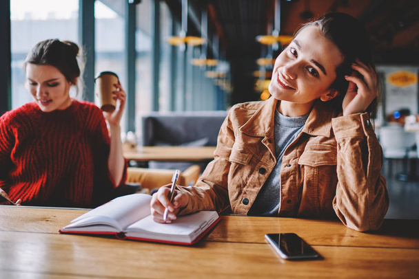Portrait of positive smiling hipster girl writing essay with excited idea in notebook while female friend sitting near and enjoying coffee, happy woman looking at camera and doing homework indoors - Фото, изображение