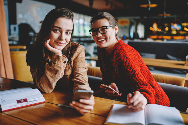 Portrait of positive female friends looking at camera while installing application for chatting using cafeteria wireless internet together, two happy hipster girls using smartphone devices indoors - Foto, immagini