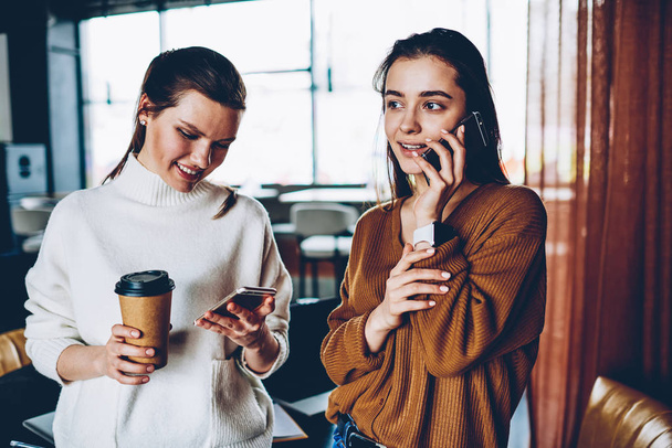 Successful hipster girl updating profile in social networks and installing app using 4g connection holding takeaway coffee while happy female friend making positive cellular call via application - 写真・画像