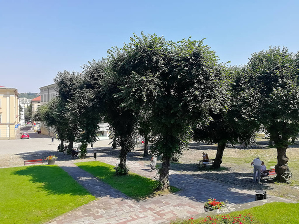 Centro da cidade em Przemysl com árvores, casas, bancos e grama bem tratada com pessoas de fundo
 - Foto, Imagem