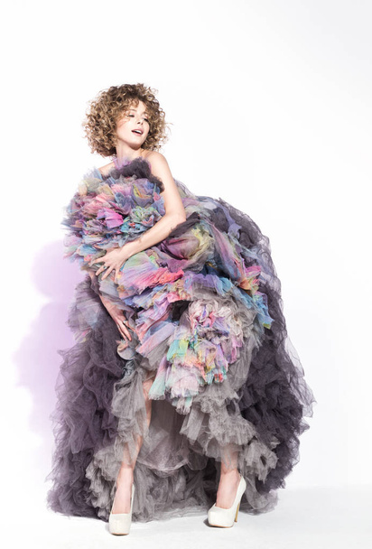 Cheerful young woman in voluminous colorful dress posing against white walls. Hairstyle with curls and a bright smile - Photo, image
