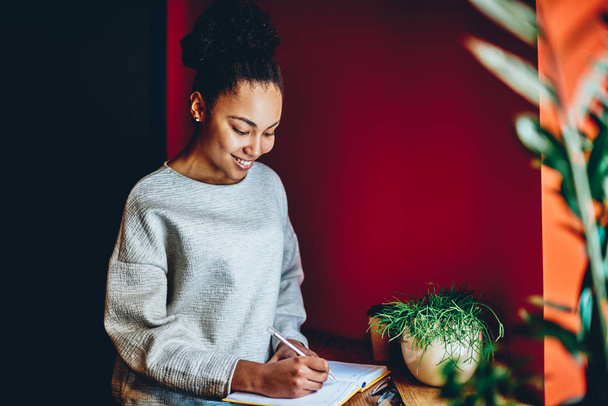 Smiling african american woman noting plannings in notepad sitting indoors in home interior, positive dark skinned hipster girl satisfied with having creative idea for publication writing in noteboo - 写真・画像