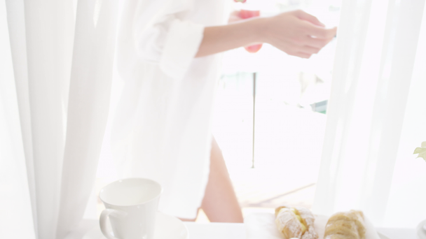 Slow Motion bliska DOF: młoda kaukaski kobieta w białej koszulce zbierając świeżo wyciszony sok owocowy na śniadanie w przepięknym pokoju hotelowym i przechodząc na taras z widokiem na żaglówkę portu - Materiał filmowy, wideo