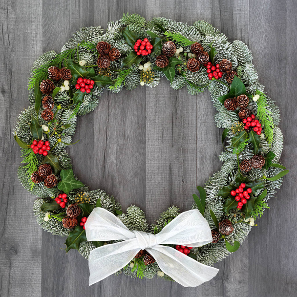 Traditional winter and Christmas natural wreath with bow, holly, snow covered spruce pine fir, mistletoe, pine cones, cedar and ivy leaves on rustic grey wood  background. - Photo, Image