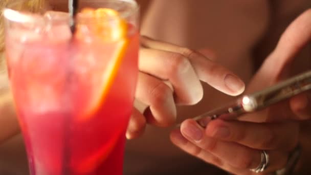 A beautiful woman with a smartphone in her hand drinks a cocktail at the bar - Imágenes, Vídeo