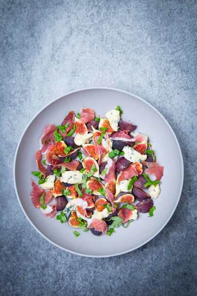 A salad comprising quartered figs with parma ham sliced and chunks of blue cheese with basil in a large grey salad bowl on a grey grunge background. - Foto, immagini